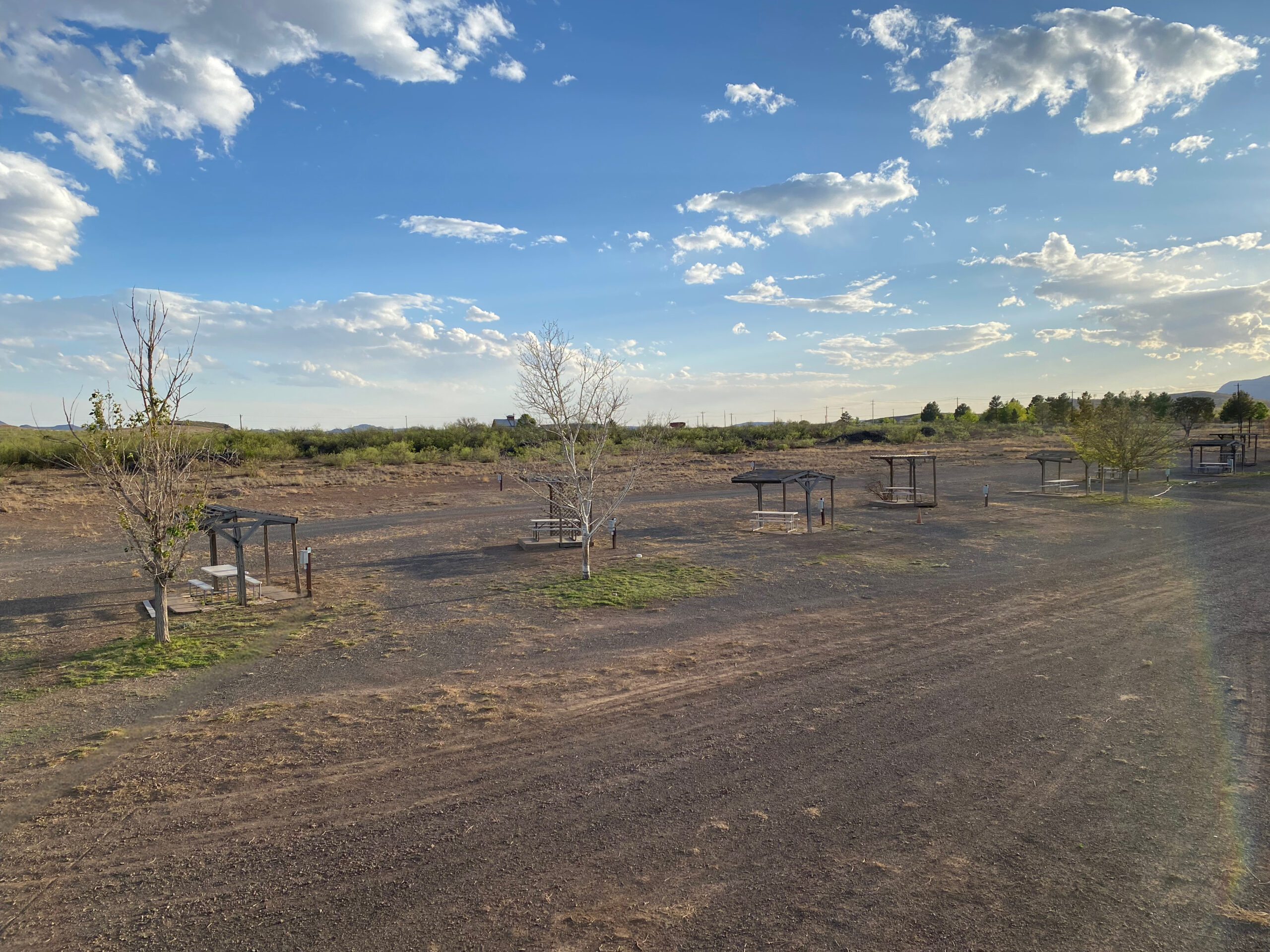 A dry, open landscape with scattered picnic tables and small covered shelters, sparse trees, patches of grass, and a dirt ground under a blue sky with scattered clouds.