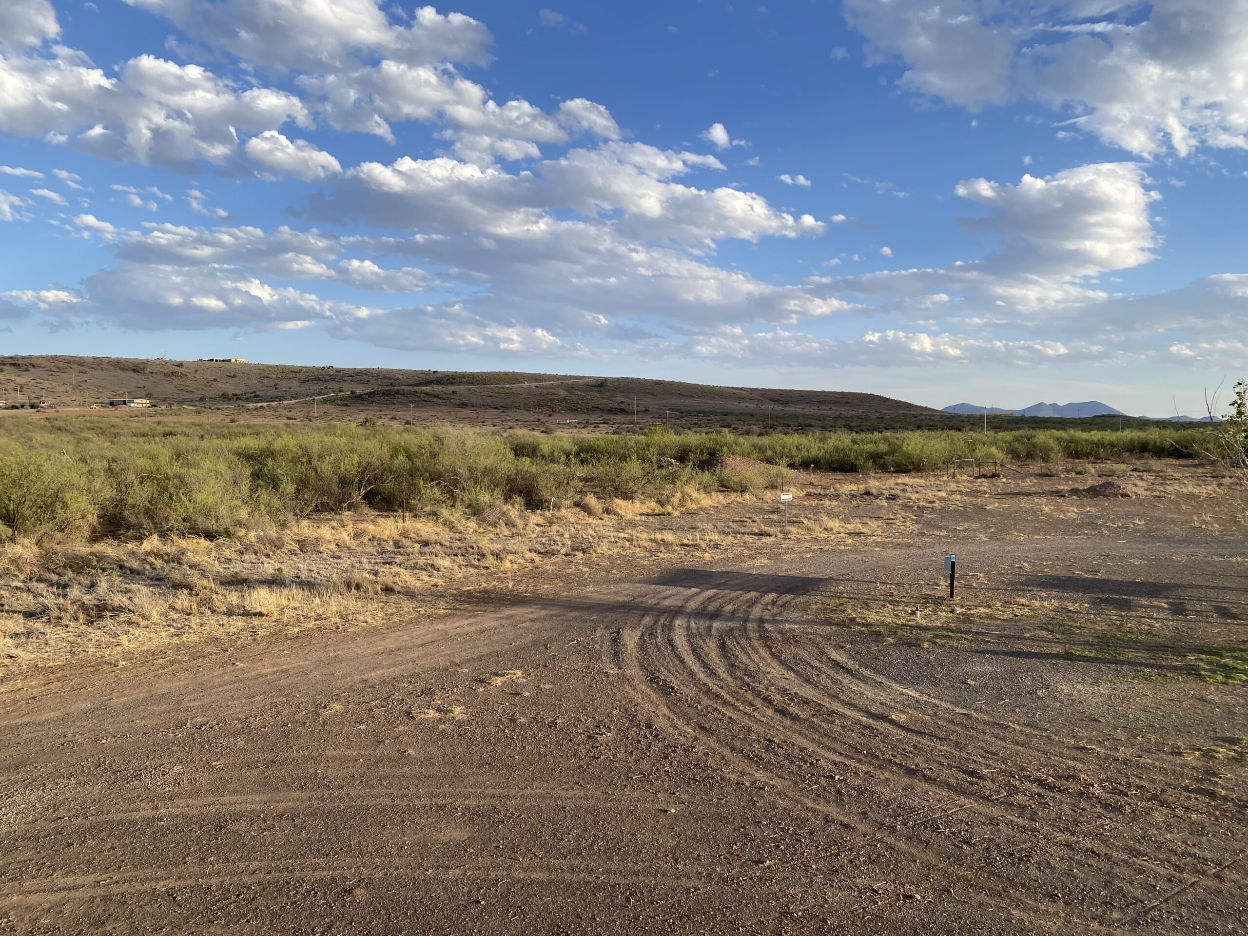 A dirt road curves through dry, grassy terrain under a partly cloudy blue sky, with shrubs and low hills in the background. The sun casts long shadows on the landscape.